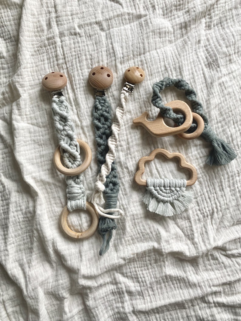 K&ouml;nnte beinhalten: Vier Holz-Bei&szlig;ringe mit Makramee-B&auml;ndern und Clips. Die Bei&szlig;ringe sind in Form eines Rings, einer Wolke, eines Wals und einer Spirale. Die Bei&szlig;ringe sind in Graut&ouml;nen und Wei&szlig;.