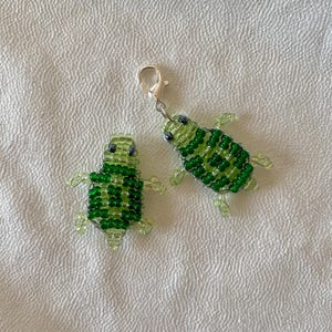 Könnte beinhalten: Zwei Schildkröten-Anhänger aus Perlen. Die Schildkröten bestehen aus grünen Perlen mit schwarzen Augen und transparenten Beinen. Ein Anhänger hat einen silbernen Verschluss. Die Anhänger werden auf einer weißen, strukturierten Oberfläche präsentiert.