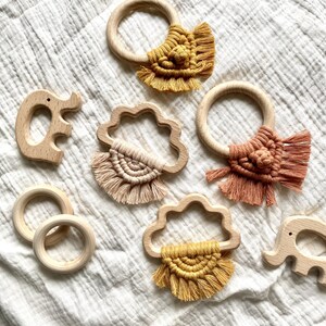 K&ouml;nnte beinhalten: Ein Set aus f&uuml;nf Holz-Bei&szlig;ringen mit bunten Makramee-Quasten. Die Bei&szlig;ringe sind in Form einer Wolke, eines Kreises und eines Elefanten. Die Quasten sind in Gelb-, Orange- und Rosat&ouml;nen gehalten.