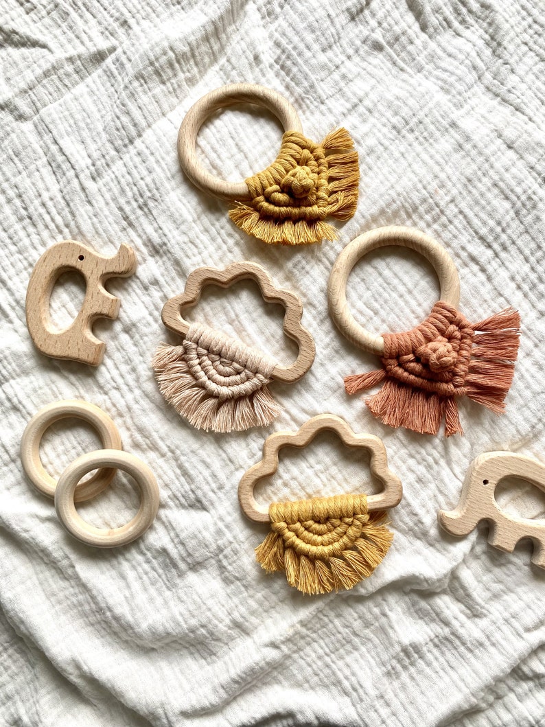 K&ouml;nnte beinhalten: Ein Set aus f&uuml;nf Holz-Bei&szlig;ringen mit bunten Makramee-Quasten. Die Bei&szlig;ringe sind in Form einer Wolke, eines Kreises und eines Elefanten. Die Quasten sind in Gelb-, Orange- und Rosat&ouml;nen gehalten.