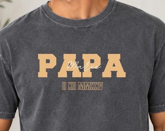 Könnte beinhalten: Ein anthrazitfarbenes Kurzarm-T-Shirt mit dem Wort PAPA in großen, hellorangen Blockbuchstaben. Unter dem Wort PAPA steht der Text II XII MMXXV in einer kleineren, passenden hellorangen Schriftart.