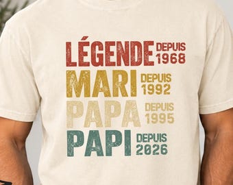 Könnte beinhalten: Ein elfenbeinfarbenes Kurzarm-T-Shirt mit gestapeltem Text in Rot, Gelb, Beige und Grün. Der Text lautet LÉGENDE DEPUIS 1968, MARI DEPUIS 1992, PAPA DEPUIS 1995 und PAPI DEPUIS 2026, mit einem abgenutzten, vintage-artigen Grafikdruck.