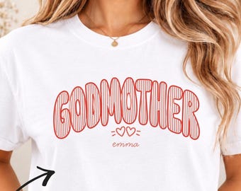Könnte beinhalten: Ein weißes kurzärmeliges T-Shirt mit dem roten Schriftzug GODMOTHER in einer gestreiften Schriftart, zwei kleinen Herzen unter dem Wort und dem Namen emma darunter. Ein schwarzer Pfeil zeigt auf das T-Shirt mit dem Text Custom Name.