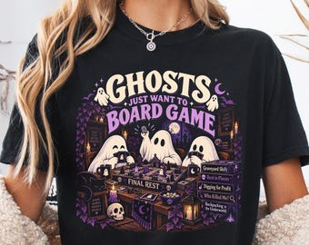 Könnte beinhalten: Ein schwarzes T-Shirt mit einem Grafikdruck von drei weißen Geistern, die ein Brettspiel namens Final Rest spielen. Das Design enthält violetten und weißen Text mit der Aufschrift Ghosts Just Want To Board Game sowie Abbildungen eines Schädels, Kerzen und Büchern.