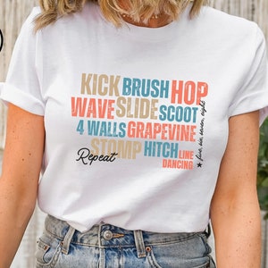 以下が含まれることがあります： ラインダンスのグラフィックがプリントされた白いTシャツ。グラフィックには「Kick Brush Hop Wave Slide Scoot 4 Walls Grapevine Stomp Hitch Line Dancing」の文字が書かれています。
