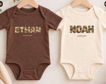 Könnte beinhalten: Zwei kurzärmelige Babybodys an Holzkleiderbügeln. Der braune Body zeigt den Namen ETHAN in Camouflage-Druck mit dem Text everson darunter. Der naturfarbene Body zeigt den Namen NOAH in Camouflage-Druck mit dem Text johnson darunter.