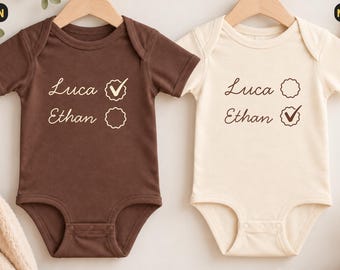 Könnte beinhalten: Zwei kurzärmelige Babybodys, einer in Braun und einer in Naturcreme. Beide zeigen die Namen Luca und Ethan in Schreibschrift mit einem kreisförmigen Kästchen daneben. Der braune Body hat ein Häkchen bei Luca, der naturfarbene Body hat ein Häkchen bei Ethan.