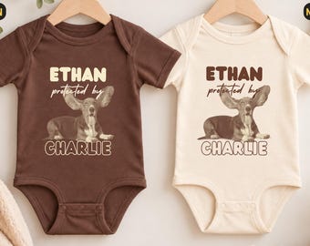 Könnte beinhalten: Zwei kurzärmelige Babybodys, einer in Braun und einer in Naturcreme, jeweils mit einer Grafik eines Dackels mit großen Ohren und dem Text ETHAN protected by CHARLIE, der in einer fetten, abgerundeten Schrift auf der Brust gedruckt ist.