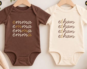 Könnte beinhalten: Zwei kurzärmelige Babybodys an Holzkleiderbügeln. Der linke Body ist braun und mit dem Namen "emma" viermal in Gold und Weiß bedruckt. Der rechte Body ist naturfarben-creme und mit dem Namen "ethan" viermal in Braun bedruckt.