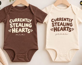 Könnte beinhalten: Zwei kurzärmelige Babybodys, einer in Braun und einer in Naturcreme, jeweils mit dem Text "CURRENTLY STEALING HEARTS" in einer fetten, abgerundeten Schrift und kleinen Herzsymbolen. Der braune Body ist mit "Mia" und der naturfarbene mit "James" personalisiert.