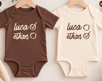 Könnte beinhalten: Zwei kurzärmelige Babybodys an Holzkleiderbügeln. Der braune Body zeigt den Text 'luca' mit einem Häkchen und 'ethan' mit einem Kreis. Der naturfarbene Body zeigt den Text 'luca' mit einem Kreis und 'ethan' mit einem Häkchen.