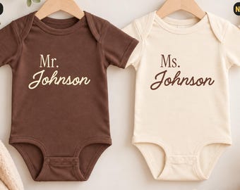 Könnte beinhalten: Zwei kurzärmelige Babybodys an Holzkleiderbügeln. Der braune Body ist mit dem weißen Schriftzug "Mr. Johnson" bedruckt, der naturfarbene Body mit dem braunen Schriftzug "Ms. Johnson". Beide Kleidungsstücke haben einen Rundhalsausschnitt.