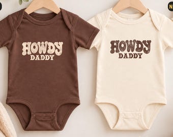 Könnte beinhalten: Zwei kurzärmelige Babybodys an Holzkleiderbügeln. Einer ist dunkelbraun mit dem Text HOWDY DADDY in hellbeiger Schrift. Der andere ist naturfarben-creme mit dem Text HOWDY DADDY in dunkelbrauner Schrift. Beide haben einen runden Halsausschnitt.
