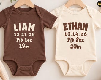 Könnte beinhalten: Zwei kurzärmelige Babybodys an Holzkleiderbügeln. Der braune Body zeigt den Namen LIAM, das Datum 12.21.26, das Gewicht 7lb 1oz und die Größe 19in. Der naturfarbene Body zeigt ETHAN, 10.14.26, 7lb 8oz und 20in.