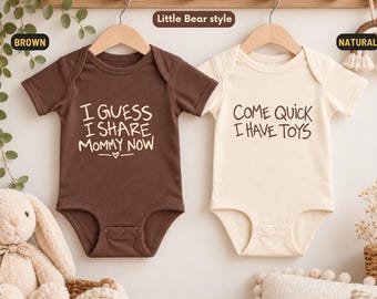 Könnte beinhalten: Zwei kurzärmelige Babybodys an Holzkleiderbügeln. Der braune Body trägt den weißen Schriftzug I GUESS I SHARE MOMMY NOW. Der naturfarbene Body trägt den schwarzen Schriftzug COME QUICK I HAVE TOYS.