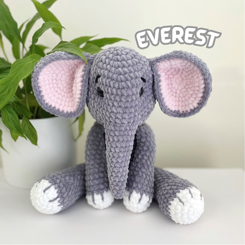 Puede incluir: Un juguete de elefante de crochet gris con orejas rosas y pies blancos. El juguete est&aacute; sentado en una superficie blanca y tiene la palabra "EVEREST" escrita en negro encima.