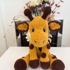 May include: A crocheted giraffe toy in shades of yellow and brown. The giraffe has a textured body, brown spots, and dark brown details on its ears, horns, and feet. It is positioned on a white surface, with a floral arrangement in the background.