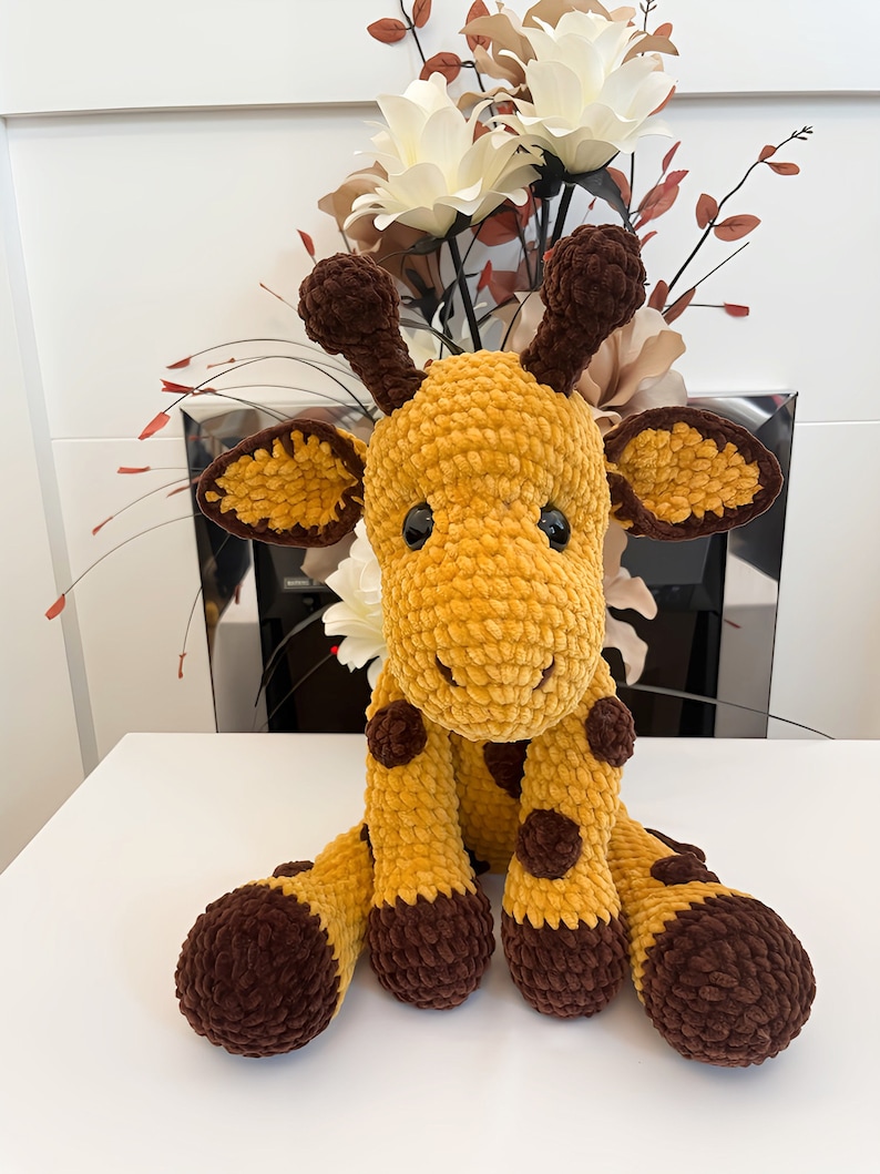 May include: A crocheted giraffe toy in shades of yellow and brown. The giraffe has a textured body, brown spots, and dark brown details on its ears, horns, and feet. It is positioned on a white surface, with a floral arrangement in the background.