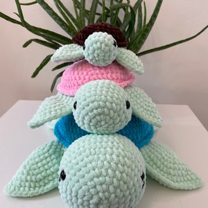 May include: A stack of three crocheted turtle toys. The bottom turtle is light green, the middle turtle is light blue, and the top turtle is pink with a brown shell.