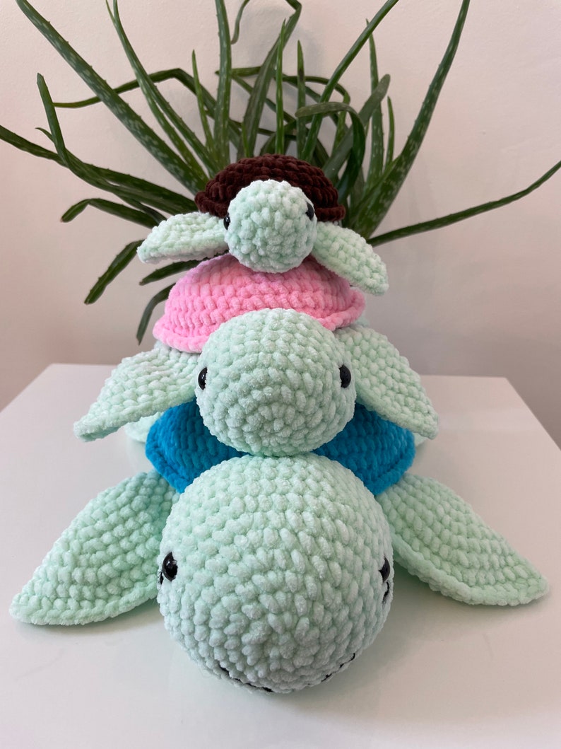 May include: A stack of three crocheted turtle toys. The bottom turtle is light green, the middle turtle is light blue, and the top turtle is pink with a brown shell.