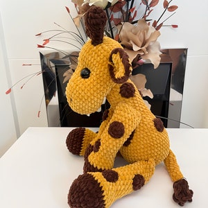 May include: A crocheted giraffe toy in yellow and brown. The giraffe has a long neck, brown spots, and a small tail. The toy is made of a soft, textured material and has black button eyes. The giraffe is a cuddly toy.