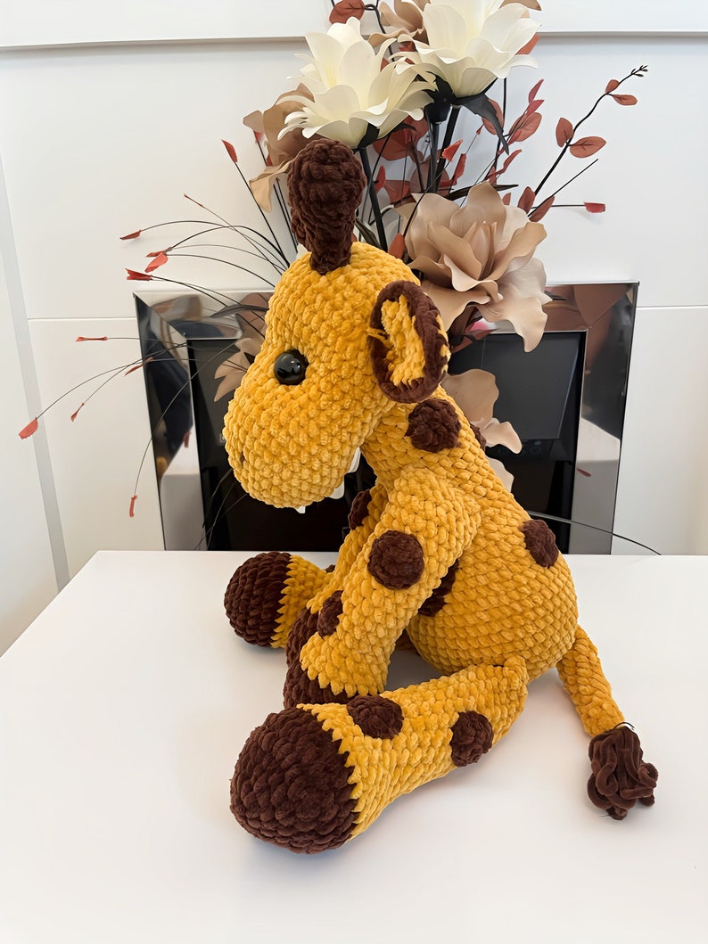 May include: A crocheted giraffe toy in yellow and brown. The giraffe has a long neck, brown spots, and a small tail. The toy is made of a soft, textured material and has black button eyes. The giraffe is a cuddly toy.