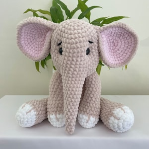Puede incluir: Un juguete de peluche de elefante tejido a crochet con orejas rosas, pies blancos y ojos negros. El elefante est&aacute; sentado sobre una superficie blanca.