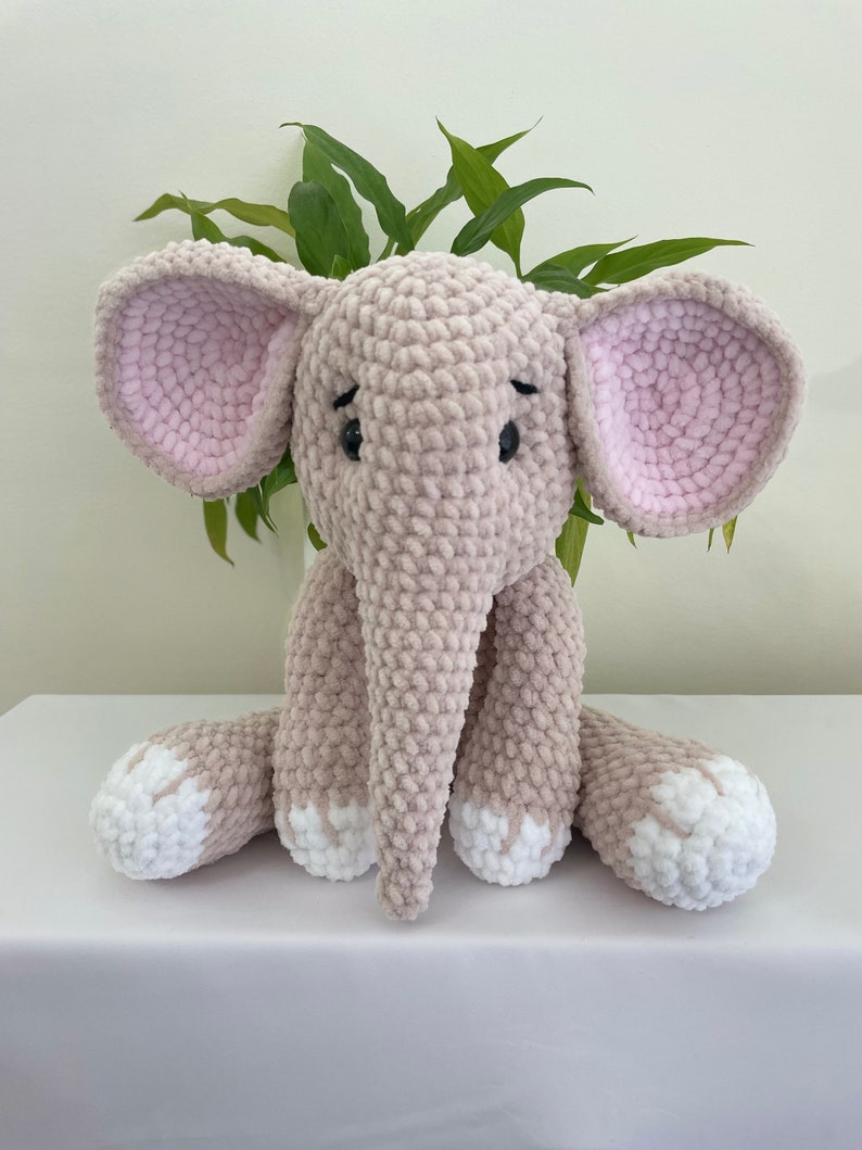 Puede incluir: Un juguete de peluche de elefante tejido a crochet con orejas rosas, pies blancos y ojos negros. El elefante est&aacute; sentado sobre una superficie blanca.