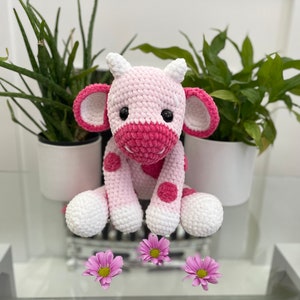 May include: A handmade crochet cow plushie in shades of pink and white, with dark pink accents on the ears, nose, and spots. The cow has large black eyes and is sitting. Pink flowers are in the foreground.