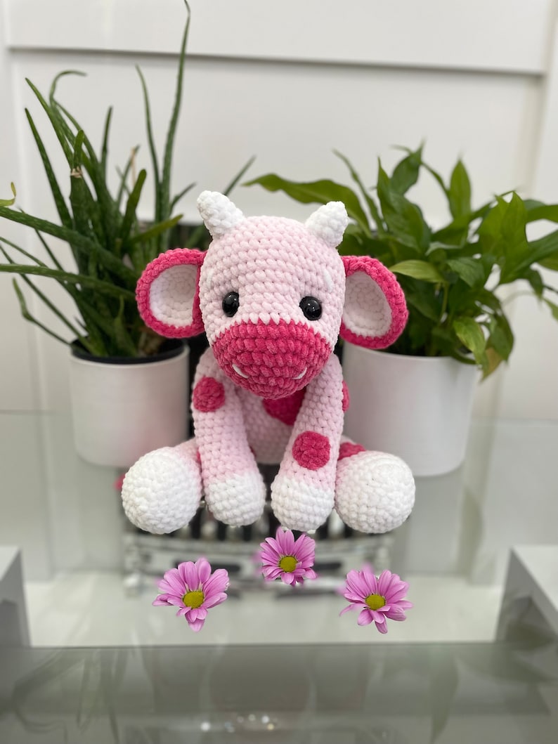 May include: A handmade crochet cow plushie in shades of pink and white, with dark pink accents on the ears, nose, and spots. The cow has large black eyes and is sitting. Pink flowers are in the foreground.
