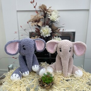 Puede incluir: Dos juguetes de elefante de crochet, uno gris y otro rosa, sentados en una cama de papel triturado con un peque&ntilde;o terrario de vidrio que contiene una planta verde en el centro.