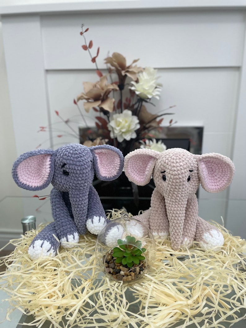 Puede incluir: Dos juguetes de elefante de crochet, uno gris y otro rosa, sentados en una cama de papel triturado con un peque&ntilde;o terrario de vidrio que contiene una planta verde en el centro.
