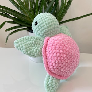 May include: A crocheted plush toy turtle with a green body and pink shell. The turtle is sitting on a white surface.