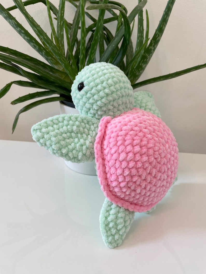 May include: A crocheted plush toy turtle with a green body and pink shell. The turtle is sitting on a white surface.