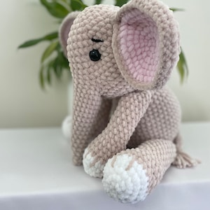 Puede incluir: Un juguete de peluche de elefante tejido a crochet con una oreja rosa, pies blancos y ojos negros. El elefante est&aacute; sentado sobre una superficie blanca.