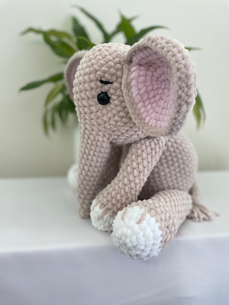 Puede incluir: Un juguete de peluche de elefante tejido a crochet con una oreja rosa, pies blancos y ojos negros. El elefante est&aacute; sentado sobre una superficie blanca.