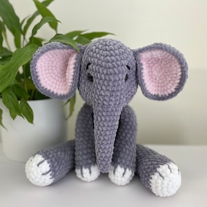 Puede incluir: Un juguete de peluche de elefante gris tejido a crochet con orejas rosas y pies blancos. El elefante est&aacute; sentado sobre una superficie blanca.