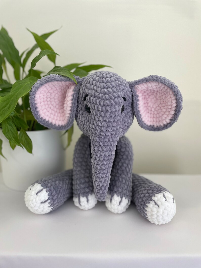 Puede incluir: Un juguete de peluche de elefante gris tejido a crochet con orejas rosas y pies blancos. El elefante est&aacute; sentado sobre una superficie blanca.