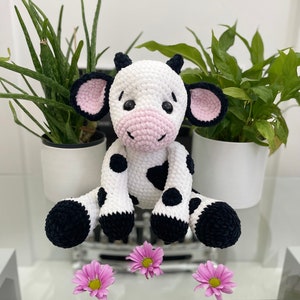 May include: A crocheted cow toy, predominantly white with black patches and hooves. It features black ears with pink interiors and a pink nose. The toy is positioned on a glass surface, with pink flowers arranged in front of it.