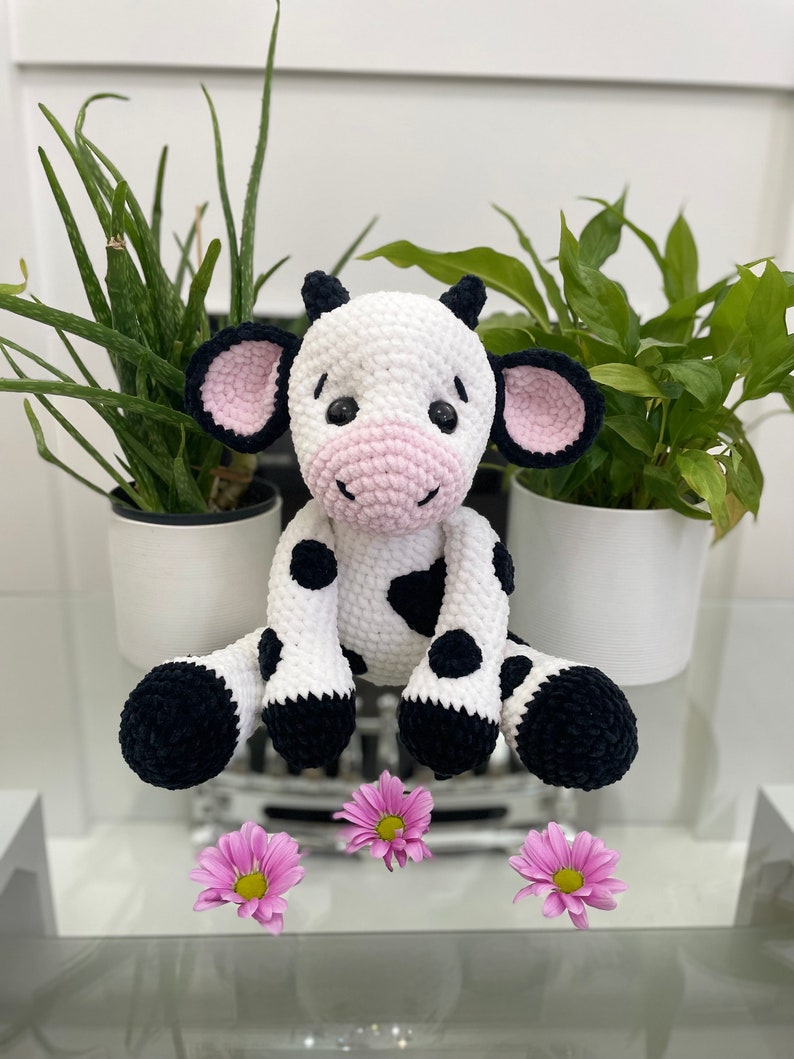 May include: A crocheted cow toy, predominantly white with black patches and hooves. It features black ears with pink interiors and a pink nose. The toy is positioned on a glass surface, with pink flowers arranged in front of it.