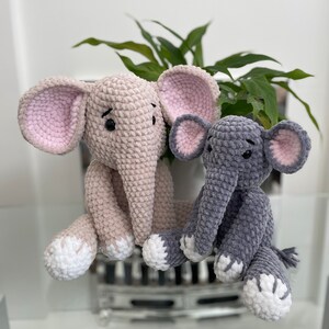 Puede incluir: Dos peluches de elefante tejidos a crochet, uno de color rosa claro y el otro de color gris. Ambos peluches tienen pies tejidos a crochet blancos y ojos negros.