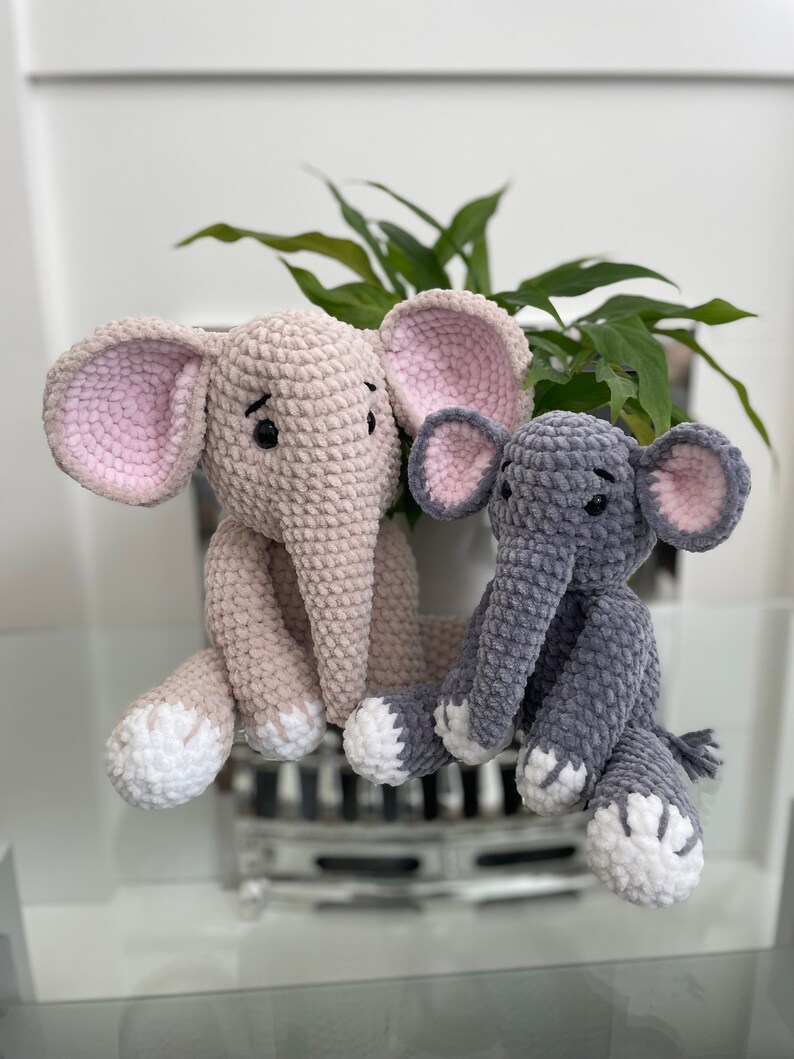 Puede incluir: Dos peluches de elefante tejidos a crochet, uno de color rosa claro y el otro de color gris. Ambos peluches tienen pies tejidos a crochet blancos y ojos negros.