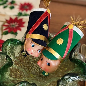 May include: Two vintage Christmas ornaments shaped like toy soldiers, one in navy blue and the other in green, with gold trim, red accents, and gold feather plumes. They rest in a green glass bowl.