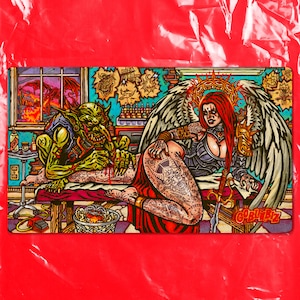 May include: A rectangular desk mat featuring a colorful illustration of a green goblin tattooing a winged figure with red hair. The scene includes a window with a dragon, tattoo equipment, and the text GOBLINBIZ in the bottom right corner.