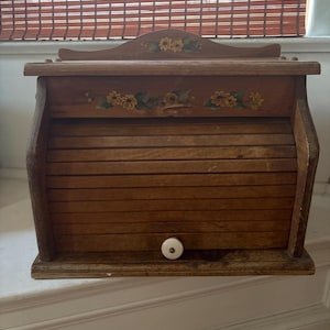 May include: A wooden bread box with a roll-top door and a white ceramic knob. The top panel and the front panel above the door feature hand-painted yellow flower designs with green leaves on a natural wood finish. The structure has a curved top.