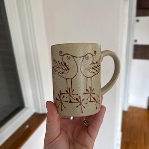 May include: A speckled beige ceramic mug featuring a brown line art illustration of two birds facing each other, standing above stylized starburst patterns. The mug has a cylindrical body and a curved handle on the right side.