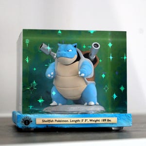 May include: A blue and tan Pokémon figure with two cannons on its back, displayed in a clear cube with a green background and star-shaped accents. The base is blue with text that reads "Shellfish Pokémon. Length: 5' 3", Weight: 189 lbs."