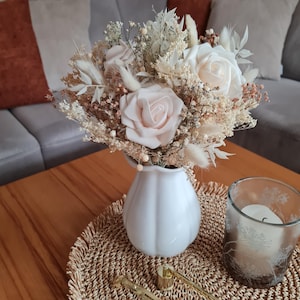 Könnte beinhalten: Eine weiße Keramikvase enthält einen Strauß aus Trockenblumen und rosafarbenen Rosen. Die Anordnung befindet sich auf einem gewebten Platzset auf einem Holztisch, mit einer Kerze in einem Glashalter. Der Strauß enthält getrocknete Gräser und kleine Blüten.