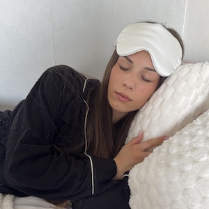 Pu&ograve; includere: Una mascherina per dormire bianca tinta unita indossata sugli occhi, abbinata a una maglia da pigiama nera a maniche lunghe con profili bianchi lungo il colletto e i bordi delle maniche. La mascherina &egrave; liscia e opaca, in contrasto con il tessuto.