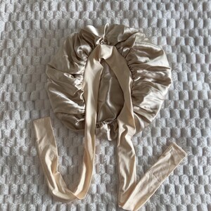 Pu&ograve; includere: Una cuffia per capelli in raso color champagne caratterizzata da un design circolare arricciato con una finitura liscia e lucente. La cuffia include due lunghi nastri in tessuto piatto che partono dal centro per una vestibilit&agrave; regolabile.